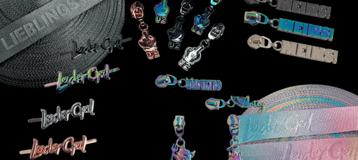 Schimmernde Metall-Zipper-Anhänger und -Zugschlaufen mit „leder-girl“-Branding in verschiedenen Farben, darunter Pink, Silber, Blau und Regenbogenverläufe.